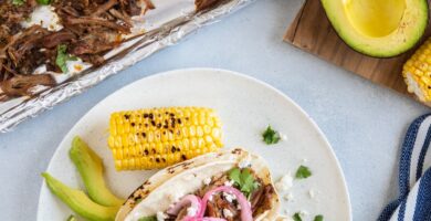 Carnitas de cerdo en una bandeja para el horno y dos tacos en un plato servidos con cebolla curtida, aguacate y maíz.